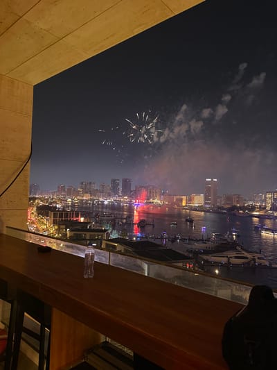 Fireworks explode in the night sky over a cityscape reflected in a waterway. Boats populate the river alongside buildings illuminated with festive lights. The view is from a high vantage point, possibly a balcony or rooftop bar, with a wooden railing in the foreground. A partial view of the building's structure is visible on the left side.