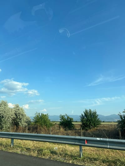 This is a vertical, eye-level, medium shot of a landscape taken from a moving vehicle, likely a car, in Tsalapitsa, Bulgaria. The primary subjects are the natural elements of the scene: a bright blue sky with scattered wispy white clouds, a line of trees and vegetation, and distant mountains. A metal guardrail runs diagonally across the right side of the frame, separating the road from the grassy verge and fields beyond.
The time of day appears to be midday, given the bright sunlight and clear sky. The weather is sunny and fair. The scene depicts a rural or semi-rural setting, possibly along a roadside. The presence of the guardrail and what appears to be a road surface on the far right edge suggests travel. The foliage is green and lush, indicating a temperate season, likely summer or spring. The distant mountains are hazy, suggesting they are some distance away.
Notable details include a small red reflective marker attached to the guardrail, and a white chalk mark that appears to be the number "6" also on the guardrail. Reflections are visible on the left side of the image, likely from the car's windows, obscuring some of the interior. There are no people or animals visible. The overall mood is peaceful and serene, capturing a moment of travel through a scenic landscape.