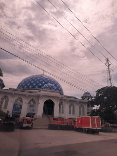 The image presents an exterior view of a large mosque and its surroundings under an overcast, cloudy sky, likely taken from a vehicle on a street. Numerous power lines stretch across the upper part of the frame.

The central subject is a grand white mosque featuring a large main dome and several smaller domes, all adorned with a distinctive blue and white diamond checkered pattern. The mosque's facade is predominantly white, characterized by multiple arched windows filled with intricate lattice designs. A wide set of stairs leads to the main entrance.

In the foreground, a dark-colored car is partially visible on the left. A small food cart is parked directly in front of the mosque, followed by a red box truck on the road. Another smaller motorized vehicle is seen further to the right. To the right of the mosque, a tall cellular communication tower stands, partially obscured by a large, dark green tree.

Several banners are affixed to the mosque's facade. One prominent banner on the left reads "Shalat Idul Adha" (Eid al-Adha prayer). Another banner to its right displays "PENGAJIAN MAGRIB JUM'AT" (Friday Maghrib Study/Recitation). A sign on the lower wall of the mosque states "MASJID ISLAMIC CENTER AL-TAQWA KAB. PIDIE JAYA." The food cart has text indicating items like "BAKSO," "SOT," "KOPI," and "ES TEH" (meatball soup, soto, coffee, and iced tea).

The scene depicts a daytime setting with diffuse light from the cloudy conditions, typical of a public space in Paya Tringgadeng, Indonesia. No direct human activity is clearly discernible, but the vehicles and food cart suggest a functional, lived-in environment around the mosque.