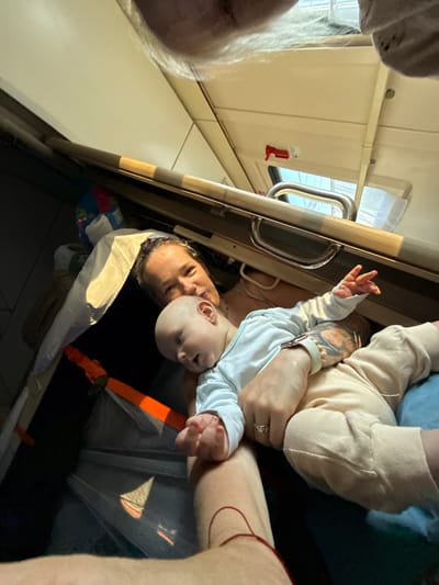 A woman is holding a baby on a train in Uzunovo, Russia. The woman is smiling and looking at the camera. The baby is looking away from the camera and is wearing a white long-sleeved shirt. The train compartment appears to be a sleeper car with beds stacked above. There are bags and luggage in the background. The lighting is dim, with sunlight coming through a window.