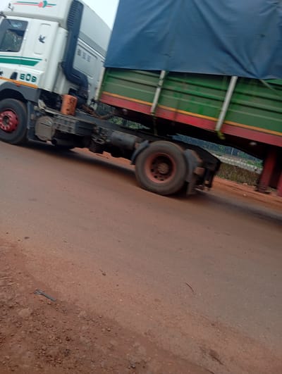 A white and green truck with a dark blue tarp-covered trailer is driving on a dirt road in Imilike, Nigeria. The truck has the number "609" visible on its side. The trailer is green with red and yellow stripes. There is a black exhaust pipe on the side of the truck. The road surface is reddish-brown dirt. Vegetation is visible in the background, along with a low brick wall. A fork and a centipede are also visible on the road. The image is blurry, indicating movement.