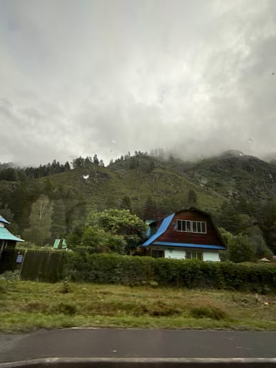 A cloudy, overcast day hangs over a mountainous landscape in the city of Uznezya, Russia. In the foreground, a paved road runs horizontally across the bottom of the frame. Behind the road, a grassy slope leads up to a dense line of green bushes. Beyond the bushes, a light blue and white house with a reddish-brown, steeply pitched roof is visible. The house has multiple windows on its upper level. To the left of this house, a smaller, dark green structure with a turquoise roof is partially visible. Further back, the landscape rises into a steep, green mountain covered in trees, with mist and clouds obscuring the upper reaches. The overall atmosphere is one of a rural, possibly mountainous, settlement under a subdued sky. No people are visible in the image.