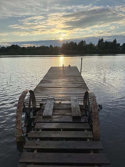 An eye-level, full shot captures a weathered wooden pier extending into a calm body of water, likely a lake or river, near Sitno, Russia. The sun, partially obscured by clouds, casts a warm glow across the scene, creating a shimmering reflection on the water's surface. Tall, dark trees line the opposite bank, silhouetted against the sky. The pier itself appears aged, with worn wooden planks and two large, rusty metal wheels situated on either side of its entrance. The water is slightly rippled, and reeds or tall grass poke out from beneath its surface. The overall atmosphere is serene and tranquil, bathed in the soft light of either sunrise or sunset. There are no people or animals visible in the frame.