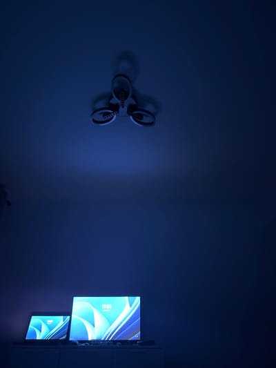 The image depicts a dimly lit room with blue ambient lighting. Dominating the ceiling is a three-pronged light fixture, casting a soft glow downwards. Below, on a dark cabinet, sit two electronic devices: a laptop on the left and a larger monitor on the right. Both screens display the same abstract wallpaper featuring flowing blue and white shapes, with the time "11:01" prominently displayed. The overall atmosphere is minimalist and modern, with no visible people or clutter. The scene is set in an indoor environment, likely a home or office. The location in Resilovo, Bulgaria, is not visually represented in the image itself, but is provided as context for the scene.