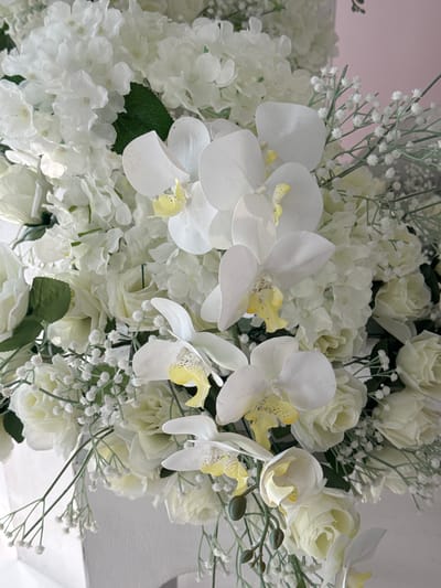 A close-up, eye-level shot showcases a bouquet of white artificial flowers, predominantly orchids and roses, interspersed with delicate baby's breath and greenery. The orchids feature prominent white petals with yellow centers, while the roses are tightly budded and layered. The floral arrangement is set against a soft pink background. No people or actions are depicted. The environment appears to be indoors, with a white rectangular object visible at the bottom, suggesting it may be a vase or part of a larger display. The specific location in Tanjungmeru, Indonesia, cannot be determined from the visual information provided.