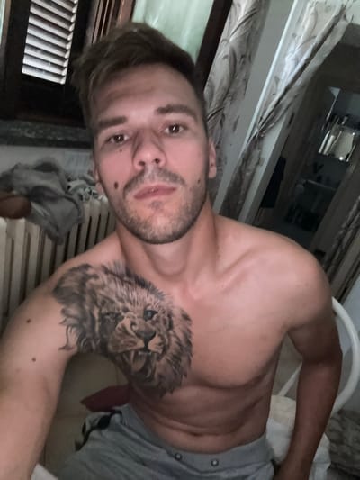 An individual with short brown hair and stubble is captured in a medium shot from the chest up, looking directly at the camera. The person is shirtless, revealing a prominent tattoo of a lion's head on their left pectoral and shoulder. They are wearing grey shorts. The background suggests an indoor setting, possibly a bedroom or living area, with a window covered by horizontal blinds on the left and draped curtains on the right. A radiator is visible behind the person on the left. The overall lighting is dim. The image appears to be a selfie taken within a residential space in Sesto Calende, Italy.