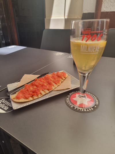 The image shows a close-up of a table with a glass of pale yellow liquid, likely beer, and a slice of bread topped with crushed tomatoes. The glass has red lettering that appears to read "1906 MIL NUEVE." It is resting on a coaster with a star logo and the words "ESTRELLA GALICIA" and "CERVEZA." The bread is served on a striped plate, with a small tan napkin tucked underneath. The table surface is dark and appears to be in an indoor setting, possibly a bar or restaurant. The lighting suggests it could be daytime or evening. The overall scene conveys a simple, casual meal or snack.