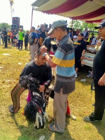 The image depicts an outdoor cultural or ritualistic event in Celep, Indonesia. In the foreground, a young adult male, dressed in a black long-sleeved shirt and dark pants, kneels on dry, yellowish-green grass. His face bears dark smudges, and he clutches a black, horse-shaped "kuda lumping" prop featuring a black mane/tail and red/black patterns on its body. An older male, wearing a grey baseball cap, a striped polo shirt (dark grey, light blue, yellow, dark grey), khaki pants, and blue flip-flops, stands over the kneeling man, gently touching his forehead with both hands. A silver watch is visible on the older man's left wrist. To the right, a third male in a black cap and shirt partially observes the interaction.
In the midground and background, a diverse crowd of approximately 20-30 individuals observes the scene. Some crowd members hold smartphones, seemingly recording. A large red and white striped canopy provides shade above the crowd. On the far left, two large black speakers on stands are visible, along with a light-colored flag on a pole. A white vehicle or trailer with a percussion instrument, possibly a kendang drum, is partially visible behind the crowd on the right. The scene is illuminated by bright daylight under a clear sky.