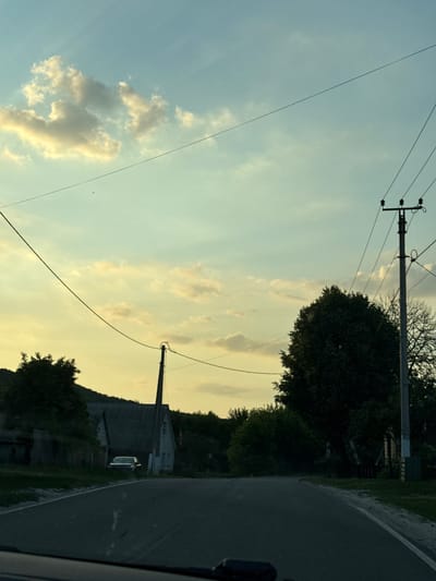 The image depicts a road curving to the right, bordered by a grassy shoulder and dark green trees. On the left side of the road, a large, dark building with a sloped roof is visible, accompanied by another smaller building. A white car is parked to the right of the larger building. Further left, power lines stretch across a pale, cloudy sky. A telephone pole with insulators is also prominent in the sky. The scene appears to be set in Nizhne-Chufichevo, Russia. The lighting suggests it is either early morning or late afternoon, with the sky illuminated by soft, yellowish light. The clouds are scattered and wispy, indicating fair weather. There are no people visible in the image, and no text is present. The overall mood is peaceful and rural.