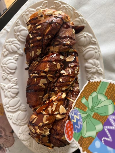 A braided loaf of bread, topped with almonds and chocolate drizzle, sits on an oval, white serving platter embossed with a pumpkin design. An Easter-themed cake topper with a green bow and Easter eggs decorates the loaf. The setting is an indoor environment, likely a home, indicated by the tablecloth beneath the platter.