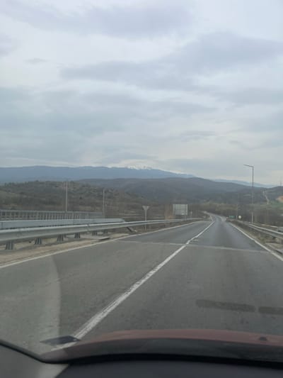 The image displays a roadside view in Marikostinovo, Bulgaria, captured from inside a vehicle, with a dark red dashboard visible at the bottom of the frame. A two-lane asphalt road, marked by a continuous white line on the right shoulder and a dashed white line in its center, stretches into the distance, gradually curving to the right. Patches of the road surface appear wet or damp.
On the immediate left, a multi-tiered metal guardrail system parallels the road, with concrete support structures visible underneath. Behind this, two tall, thin utility poles stand. Beyond the guardrail, another road or structure runs parallel to the main thoroughfare. On the right side of the main road, a single metal guardrail is present. Further ahead on the right, a white triangular yield sign with a red border is visible, followed by a grey rectangular road sign and a tall, dark street light pole.
The mid-ground features rolling hills covered in sparse, bare deciduous vegetation, indicative of a late winter or early spring landscape. In the distant background, a prominent mountain range extends across the horizon, with several peaks exhibiting patches of white snow. The sky above is entirely overcast with numerous light grey clouds, resulting in diffused ambient lighting across the scene. No individuals, animals, or active interactions are depicted.