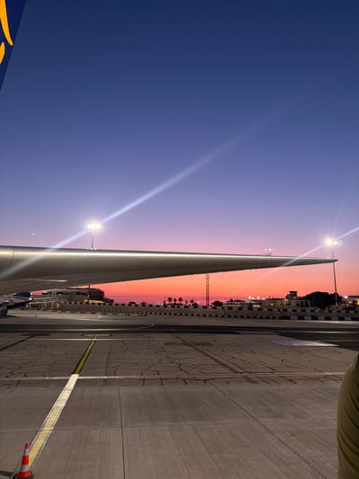 The image captures an airport scene in Luqa, Malta, at dusk. The sky transitions from deep blue to orange. The wing of a parked aircraft dominates the left side of the frame. The airport apron, marked with yellow lines, stretches into the right side of the image. Several streetlights illuminate the area and cast glares. A red and white checkered wall runs along the edge of the apron, bordering buildings and structures.