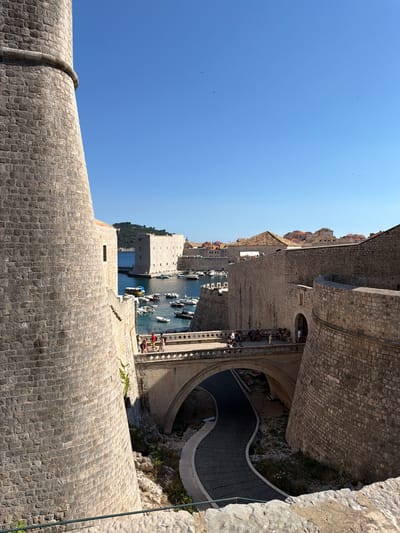 The image captures a view from the city walls of Dubrovnik, Croatia, looking down towards the harbor. The sky is a clear, bright blue, indicating a sunny day, likely in the late morning or early afternoon given the strong sunlight casting shadows. The ancient stone walls, constructed from rough-hewn blocks, dominate the foreground and midground, showcasing their impressive scale and texture.
Below the walls, a picturesque harbor is visible, filled with numerous small boats and yachts moored in the clear blue water. In the distance, more buildings with terracotta roofs, characteristic of Dubrovnik's Old Town, are nestled against a backdrop of lush green hills. A curved, paved pathway with railings snakes down from the wall, and several people can be seen walking on it or sitting on the steps, enjoying the view.
The scene is one of historical grandeur and vibrant activity. The sturdy stone fortifications suggest the city's rich past, while the bustling harbor and the presence of visitors point to a popular tourist destination. The atmosphere appears peaceful and serene, with the sunlight illuminating the ancient architecture and the sparkling water. No visible text is present in the image.