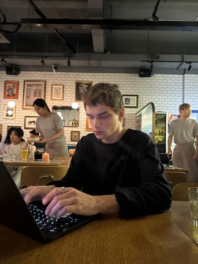 The image depicts a young man with short, light brown hair, wearing a black long-sleeved shirt and several rings on his fingers, seated at a wooden table in what appears to be a restaurant in Xuhui District, China. He is actively typing on a black laptop, which is positioned on the table in front of him. The background reveals a dimly lit restaurant interior with exposed ceiling elements and industrial lighting fixtures. White tiled walls are adorned with framed pictures and artwork. Several other individuals are present in the background: a woman in a light gray uniform is carrying a bowl, suggesting she might be a server; another woman with dark hair is seated at a table to the left, looking down; and a man in a gray shirt and apron is standing further in the background near what looks like a bar area. Glasses and a lit candle are visible on the table in the foreground. The overall atmosphere suggests a casual dining setting with patrons engaged in various activities.