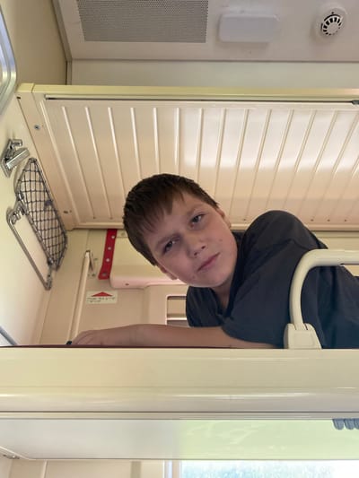 A young boy with short brown hair and blue eyes is pictured upside down, looking at the camera. He is wearing a dark gray t-shirt. The boy is inside a small, enclosed space with cream-colored, slatted walls. A white curved bar is visible behind him, suggesting he might be in a vehicle, possibly a train compartment.
To the left of the boy, a white round object with a black center, resembling a smoke detector, is mounted on the wall. Below it, a rectangular white object is also attached to the wall. Further down, a metallic grille is visible.
On the wall directly behind the boy, a red band with small circular holes runs horizontally. To the right of this band, a small sticker with text and a red triangular symbol is affixed to the wall.
In the foreground, on a cream-colored surface, a metal rack with a mesh surface is positioned. Next to it, a silver metal clip or latch is visible.
The lighting suggests it is daytime. The overall scene appears to be indoors, within a confined space. The boy's expression is neutral. The location is identified as Ershovka, Russia.