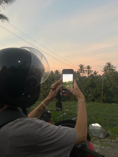 A person wearing a black helmet and a grey t-shirt is seated on a red scooter, holding a smartphone to capture a scenic view. The person's left arm is adorned with a bracelet of white shells and a tattoo of a mushroom on their forearm. The smartphone is displaying a landscape with a road and a field, framed by lush green trees and palm trees under a soft pink and blue sky, indicative of a sunset. The background features a rural landscape with agricultural fields and utility poles. This scene is set in Loloan, Indonesia.