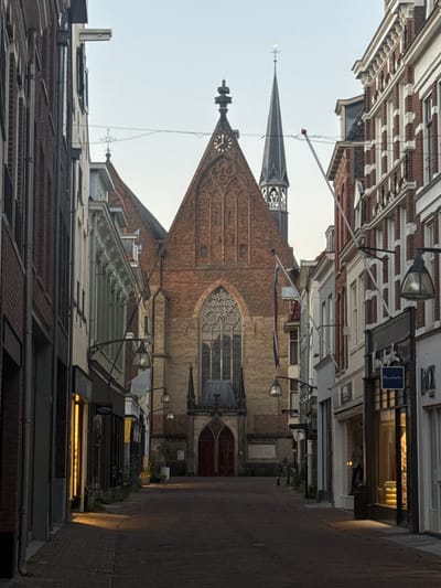 The image captures a narrow street in the city of Deventer, Netherlands, dominated by an imposing brick church structure at the end. The architecture suggests a historic European city center. On the left and right sides of the street are buildings that appear to be shops, with large display windows, some of which are illuminated, indicating the presence of businesses.
The church, the central and most prominent feature, has a large rose window above its entrance and a tall, slender spire. A clock face is visible on the church's facade. Flags hang from some of the buildings lining the street. The street itself is paved with bricks. The lighting suggests it is either early morning or late evening, with soft, diffused light and some artificial lighting from the shops.
There are no visible people or active interactions. The scene is static and appears to be an empty street, perhaps before or after business hours, or during a quiet time of day. The overall impression is one of historical depth and architectural grandeur, with a sense of calm and stillness. The specific location is likely a prominent street leading to a church in the historic center of Deventer.