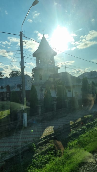 The image shows a church with a tall, slender tower topped with a cross. The sun is shining brightly in the sky, creating a glare that obscures some of the details of the church and the surrounding environment. In front of the church, there are several tall, narrow trees. A street light is visible on the left side of the image, along with power lines. The church is situated in a somewhat rural or suburban area, with houses and other buildings visible in the background. There is a fence in front of the church, and a paved area in the foreground. The overall scene suggests a peaceful, sunny day in a Romanian village.