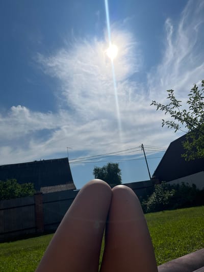 The image is a low-angle shot taken outdoors in what appears to be a residential backyard setting in Pushkino, Russia. The foreground features a person's bare legs, suggesting they are lying down or sitting, possibly on a lounge chair or the grass. The sun is bright and high in the sky, casting a lens flare across the image and creating strong highlights. The sky is a vivid blue with wispy white clouds scattered throughout, indicating clear and sunny weather.
In the mid-ground, the roof of a dark-colored building, likely a house or shed, is visible. Above this building, there are power lines and a small antenna. Further into the background, there is a well-maintained green lawn and lush green trees, some of which overhang into the frame from the top left. The overall impression is one of a pleasant, sunny day, likely during the warmer months of the year, suitable for outdoor relaxation. The cropping and focus suggest a casual, personal snapshot rather than a professional photograph. No text is visible in the image.