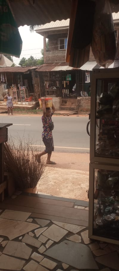 The scene depicts a street view in Umuoji, Nigeria, captured from the threshold of an establishment. In the immediate foreground, a bundle of dried reeds or grasses stands upright on the left. The ground consists of mismatched, light-colored broken tiles and a dark, horizontal wooden panel. To the right, the edge of a white-framed display case is partially visible.
Across a reddish-brown dirt shoulder and an asphalt road, a young male individual walks from right to left. He is carrying a rectangular, light-colored plastic container on his head, supported by his raised hands. He wears a multi-colored, patterned short-sleeved shirt, dark shorts with vertical white stripes, and dark sandals. A black fanny pack or sling bag is worn across his chest. Further left on the same side of the street, a female individual stands stationary on the dirt shoulder, facing towards the road, with her right hand raised to her head. She wears a white dress with a dark pattern.
In the background, a row of commercial structures lines the opposite side of the street. These include single-story buildings with corrugated metal roofs, some displaying visible rust, and open storefronts revealing various merchandise. One two-story building features unpainted cinder block construction on its lower level and a plastered upper level with a single window. Sparse green foliage is visible between and behind the buildings under a bright, overcast sky.
