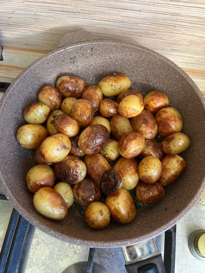 A top-down shot shows a mottled brown, non-stick frying pan filled with small, whole potatoes that have been roasted or pan-fried. The potatoes are a mix of golden-brown and lighter yellow, with some exhibiting darker, crisped skin. They are lightly seasoned with visible green herbs, likely dill, and glistening with oil. Some potatoes have a slightly wrinkled texture, indicating they are cooked through.
The pan sits on a stove top. To the left of the pan, a light-colored wooden surface with a vertical grain pattern is visible, suggesting a countertop or table. To the right of the pan, the metallic grates of a gas stove are apparent, along with part of a burner control knob, which is yellow and silver. The overall setting appears to be a kitchen.
The lighting suggests daytime, likely indoors. There are no people visible. The focus is entirely on the food and the cooking vessel. The image captures a moment of food preparation, showcasing a simple, appealing dish. No text is visible in the image.