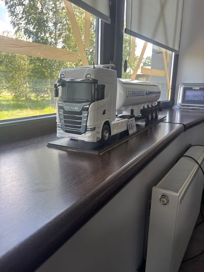 A detailed, close-up shot captures a model truck with a tanker trailer positioned on a dark, polished wooden windowsill. The model, predominantly white and gray, features the "SCANIA" logo on its front grille and "LPGPROGRESS" written on the trailer. The truck is mounted on a black display base.
The windowsill runs along the bottom of a window that looks out onto a blurred background of lush green trees and foliage under a bright sky, suggesting an outdoor environment. The window itself is partially obscured by light-colored roller blinds and strips of light brown tape applied in a diagonal pattern across the glass.
To the right of the truck model, a white radiator is visible against a gray wall. A digital display device is also present on the windowsill to the right of the truck, its screen showing faint text and numbers. The overall scene appears to be an indoor setting with a view of nature through the window. The precise location is the city of Pereiaslavske, Ukraine.