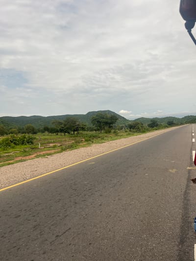 The image depicts a road in Minda, Nigeria, with a view of lush green hills and a cloudy sky. The road is asphalt with a yellow line marking the edge. The scene is likely captured during the day, given the bright sky. There are trees and vegetation along the side of the road, suggesting a rural or semi-rural setting. The perspective indicates the photo might have been taken from a moving vehicle or by someone standing by the roadside. The cloudy sky suggests it might be a humid or overcast day. There are no visible people or specific objects that stand out, other than the road itself and the natural landscape. The overall mood is tranquil, focusing on the vastness of the landscape and the open road.