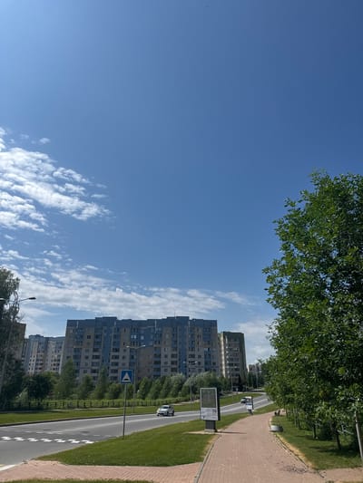 A bright, sunny day illuminates a residential district in Koltsovo, Russia. The sky is a vast expanse of clear blue, dotted with sparse white clouds. Tall, multi-story apartment buildings, painted in shades of blue and beige, dominate the background, their windows reflecting the sunlight. In the foreground, a paved walkway lines a green lawn, bordered by a line of lush green trees. A road runs alongside the lawn, with a white car visible in the distance. A pedestrian crossing is marked on the road. A signpost with a triangular symbol indicating a hazard stands near the roadside. The overall scene depicts a typical urban environment on a pleasant day.