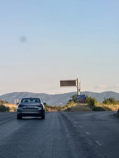 The image depicts a road scene with a car driving away from the viewer. The car is a silver sedan with a license plate reading "EB 3537 BC". The road is asphalt with faded white lane markings. On the right side of the road, there is a blue directional sign pointing towards "DEBELSKOV" and "SVILENGRAF". Further along the road, there is an electronic display board mounted on a pole. The background consists of rolling hills covered in vegetation and trees, with a clear blue sky above. A small dark speck, possibly a bird or insect, is visible in the sky on the left side of the frame. The overall impression is a rural or semi-rural environment in Bulgaria, possibly near the city of Prisovo, given the context of the directional sign. There are no people visible in the image, and the action is limited to the car driving on the road. The lighting suggests it is daytime, likely late afternoon due to the warm tones and long shadows.