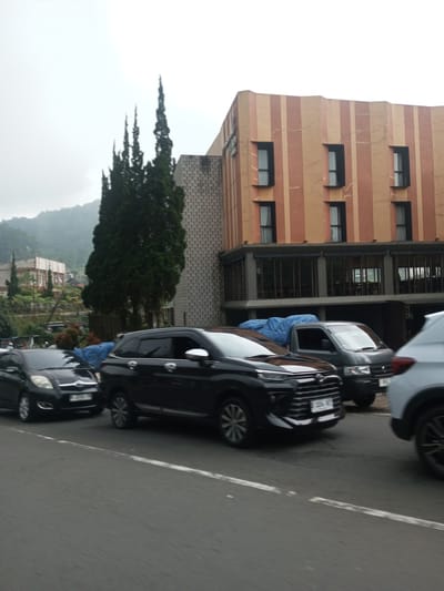 The image shows a street scene with several cars in front of a building. The building has a modern architectural style, with alternating vertical stripes of orange and beige. It has a large glass-enclosed ground floor. Tall cypress trees stand near the building. Behind the building, mountains are visible. The cars are a mix of makes and models, including a black hatchback, a dark SUV, a small truck with a blue tarp, and a white SUV. The scene takes place on a cloudy day. Based on the license plates on the car and the surrounding landscape, the scene is in the vicinity of Mae Chan District, Thailand.