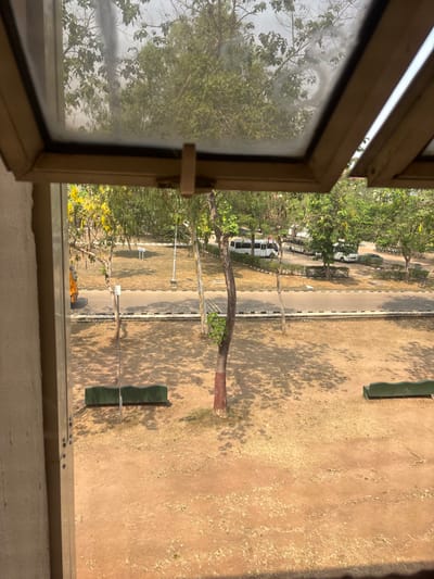 The image presents an outdoor scene in Ado-Ekiti, Nigeria, observed from an elevated window. The window frame, constructed of a light brown or beige material, is partially open, framing the view. The immediate external foreground consists of dry, reddish-brown earth with sparse, short patches of dried vegetation, suggesting arid conditions or a dry season. Multiple elongated shadows from unseen trees are cast across this ground. Two dark green, undulating benches are positioned on the dry earth, one on the left and another on the right, both casting clear shadows. Several trees with sparse foliage and varying trunk colors are present, including one central tree with a distinctive reddish-brown lower trunk.

Beyond the foreground, a paved asphalt road is visible, bordered by white and black striped curbing. No vehicles are actively in motion on this road, nor are any pedestrians present. A white utility pole or signpost stands upright near the road on the left side. The background features a cluster of vehicles parked in an open area, including at least two white passenger vans or minibuses, a white car, and a darker vehicle. This area is surrounded by numerous trees with vibrant green leaves, some exhibiting yellow blossoms. The sky is a pale, hazy blue, indicating daylight with atmospheric haze. No human figures or specific actions are discernible within the frame.