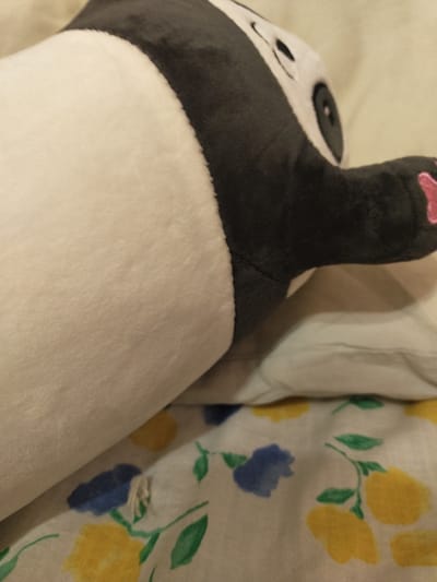 A close-up shot features a plush panda toy resting on a surface, possibly a bed. The panda is mostly white and black, with visible black ears and eye patches. Its paw, with pink toe pads, is raised. To the right of the panda, a white cloth with a floral pattern of yellow and blue flowers and green leaves is visible. The lighting suggests an indoor setting, possibly during the day. The image is taken from a high angle, focusing on the panda and the patterned cloth. No text is visible.