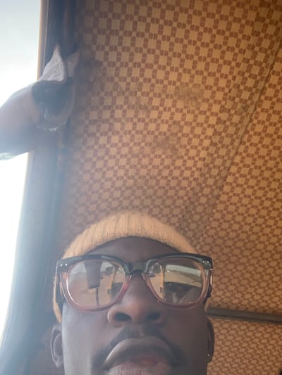 A close-up, low-angle, indoor shot shows the face of a Black individual from the nose upwards. They are wearing a light-colored beanie and thick-rimmed glasses with pink frames. The background is a textured, checkered fabric in shades of beige and brown, suggestive of a vehicle's interior. A faint light source is visible on the left side, possibly a window. The scene is set in Nabatum, Nigeria.