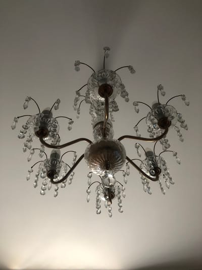 An ornate, multi-armed chandelier with clear glass crystal pendants hangs from a white ceiling. The chandelier has a metallic finish, likely brass or bronze, and features several bulbs. The glass elements reflect light and create a decorative pattern. The image was taken from below, looking up at the chandelier against a plain ceiling. There is no indication of the city of Tutaev, Russia in the image.