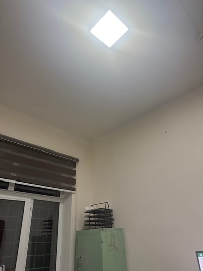 A low-angle shot reveals a room corner. On the left, a bright square ceiling light illuminates a white wall. Shifting to the right, the ceiling angles down, and a window with partially closed blinds appears. Above the window, a greenish-painted cabinet is mounted, accompanied by a dark, rack-like structure extending from its side. A small, dark spot is visible on the ceiling.