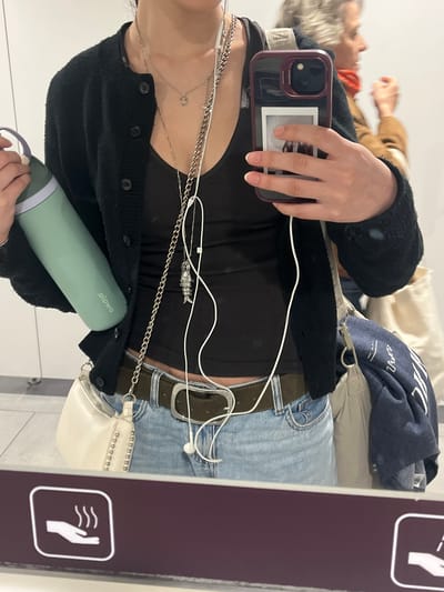 The image shows a person's selfie in a mirror. The person is wearing a black cardigan over a black top, blue jeans with a brown belt, and several silver necklaces. They are holding a cell phone in one hand, a green water bottle in the other, and wearing white earphones. There is a white purse with silver studs hanging from their belt. The reflection in the mirror shows other people in the background. In the foreground, there is a sign with a white hand over curved lines, on a purple background. It's unclear if this photo was taken in Colombier-Saugnieu, France, as no clear landmarks or signs indicating the location are visible in the image.