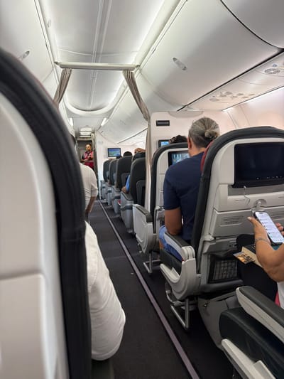 The image captures the interior of an airplane cabin, likely during flight. The perspective is from a passenger's seat, looking forward down the aisle. The scene is dimly lit, suggesting a time when cabin lights are dimmed, possibly during a long flight or a portion of it where passengers are resting.

Several people are visible, seated in the airplane's rows. In the foreground, a white pillow or headrest is prominent. Further down the aisle, a person with their hair in a bun is seated, looking at a smartphone. Another person in the distance is wearing a red garment. The seats are typical of an airplane, with tray tables and personal screens. The overall atmosphere appears calm and functional, characteristic of air travel.

There are no explicit weather cues, but the indoor setting of an airplane implies travel irrespective of external conditions. Location cues are limited to the airplane interior itself, with standard overhead compartments, air vents, and seat designs. The presence of individual screens suggests it might be a medium to long-haul flight.

Visible text is minimal, with a small label on the overhead compartment that is too small to read clearly. A tag or label is also visible on the seatback in front of the person with the bun. The image does not convey specific emotions beyond a general sense of travel. The activity is passive, with passengers engaged in personal activities like using their phones or resting.