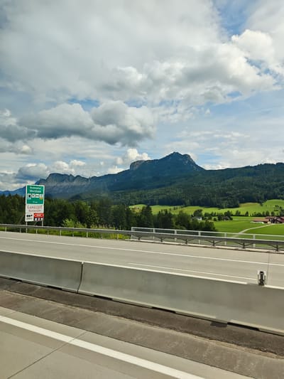 This is a medium-shot, horizontal, eye-level, outdoor, daytime photograph taken from a vehicle on a highway. The scene captures a view of a mountain landscape in St. Lorenz, Austria, on a cloudy day. In the foreground, a concrete highway barrier runs horizontally across the lower part of the frame, with a paved road extending beyond it. To the left of the road, a green sign displays "Raststation Mondsee," indicating a rest stop for Mondsee, along with icons for a gas station, toilets, and a restaurant. Below this sign, another sign reads "LANDZEIT Restaurant" with logos for OMV and VIVA. In the midground, lush green trees form a dense forest line, with rolling green fields and a few buildings visible in the distance on the right. The background is dominated by a large, dark green mountain range, with its peaks obscured by dramatic, white, and gray clouds. The sky is a mix of blue patches and heavy cloud cover.