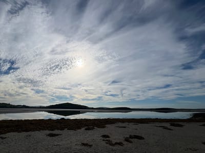 This is a scenic photograph of a tranquil beach and body of water, likely a bay or estuary, under a partly cloudy sky. The foreground features a sandy beach with a scattering of dark seaweed. Beyond the beach, a calm expanse of water reflects the sky and the distant, rolling green hills. The sky is a mix of bright blue patches and wispy white clouds, with the sun visible as a bright, diffused orb creating a subtle lens flare. The reflections on the water are clear, mirroring the landscape and clouds. The overall impression is one of peace and natural beauty, suggesting a calm, possibly cool, day, with the sun indicating it is daytime. The presence of hills and the overall landscape suggest a coastal or lakeside location in a rural area. Given the location context of Dunuisce, Ireland, this scene likely depicts a serene Irish coastal landscape. There is no visible text in the image.