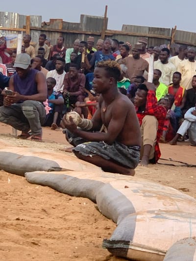 The image shows a scene in Sokoto, Nigeria (13.049, 5.238) with a large crowd of men gathered around a young man wearing jeans shorts and a black undershirt. The young man is holding a large piece of wood and looking towards the left of the image. The crowd is seated on the ground, which is covered in a reddish-brown sand. There are several large sandbags in front of the seated young man. The scene is likely a wrestling match, with the young man possibly a wrestler or a spectator. The men in the crowd are wearing a variety of clothing, with many wearing traditional garb. They are all engaged in watching the event, with some talking among themselves. The atmosphere is lively and energetic, with the men seemingly invested in the event. There is a wooden fence in the background, and a few people are standing in front of it, watching the spectacle. The image is shot from a low angle and captures the essence of the event, focusing on the young man at the center of the action. The image captures a moment of intense anticipation and engagement, giving a glimpse into the vibrant cultural practices of Sokoto, Nigeria.