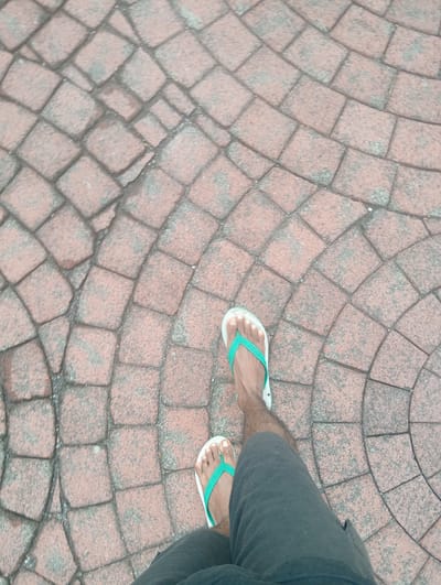 The image shows a person's feet wearing flip-flops on a brick-paved pathway. The bricks are reddish-brown and arranged in a circular pattern. The pathway appears to be outdoors, possibly in a park or public space. The lighting suggests it is daytime, but the sky is not visible. There is no indication of the specific location within Surabaya, East Java. The person is wearing dark-colored shorts.