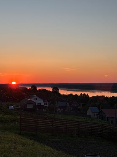 The image depicts a sunset over a rural landscape in Зеленец, Russia. The sky displays a gradient from light blue-grey at the top to vibrant orange and red closer to the horizon. The sun, an intense red-orange orb, is partially visible and low on the left horizon, casting a bright glow and reflecting off a broad body of water, likely a river, which traverses the mid-ground horizontally. The water also shows a warm, reflective surface, suggesting calm conditions.
A dense band of dark green foliage, predominantly trees, lines the banks of the water body, appearing in silhouette against the bright sky and water. On a gently sloping hillside in the lower half of the image, several residential houses are scattered among trees and green vegetation. These houses feature varying architectural styles, including structures with white walls and dark roofs, and others with darker facades and pitched roofs. Utility poles and overhead power lines are visible, crisscrossing the scene in the mid-ground, partially obscuring the houses and trees.
In the foreground, a dark wooden slatted fence extends horizontally, separating a grassy area on a slope from tilled or dark soil directly beneath it. The foreground vegetation consists of green grass and some patches of darker, disturbed earth. No people or animals are visible in the scene. The overall lighting indicates either late sunset or early sunrise.