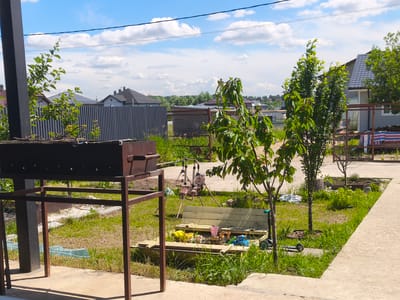 The image depicts a residential backyard scene in the village of Фронтовики Снт, Russia, under a partly cloudy blue sky. In the foreground, a rusty metal barbecue grill stands on a concrete patio. To the right of the grill, a wooden sandbox filled with children's toys, including toy trucks and buckets, is visible. A small pink swing set with a plush toy is situated behind the sandbox. Several young trees with green leaves are scattered throughout the yard. Beyond the yard, there are houses with gray roofs and a tall, vertical striped fence. The ground is covered in green grass and some dirt patches. A light-colored concrete pathway runs along the right side of the image.