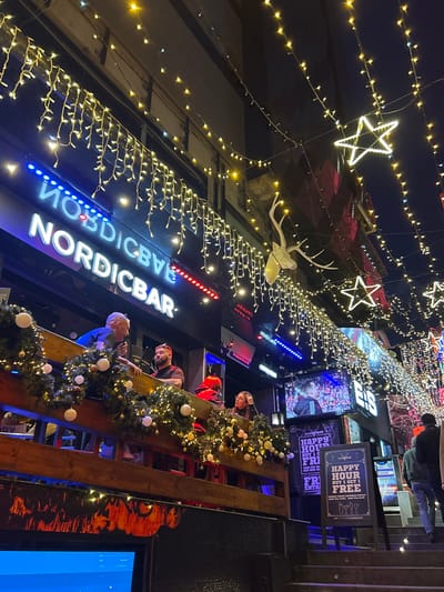 The image displays a nighttime street scene in Paceville, Saint Julian's, Malta, focused on the entrance of the "NORDICBAR" establishment. The bar's facade features an illuminated sign with "NORDICBAR" in white capital letters on a dark background, flanked by horizontal blue and red LED strips. Above the sign and across the street, numerous warm yellow LED string lights are strung, some in an icicle pattern, along with several illuminated five-pointed star ornaments. A white, stylized deer head with antlers is mounted on the building facade to the right of the bar's sign.
On the second-story balcony of "NORDICBAR," a wooden railing is decorated with evergreen garlands, white spherical ornaments, and additional string lights. Four individuals are positioned along this railing. On the left, an older male with short grey hair and a dark shirt leans over the railing. Next to him, a bearded male with a red headband and a dark t-shirt leans against the railing. To their right, a female with red hair and a red top, and another female with dark hair, also lean against the railing.
Below the balcony, to the right, a large digital screen displays a sporting event. Adjacent to this screen, a blackboard-style sign advertises "HAPPY HOUR FRI & SAT BUY 1 GET 1 FREE," with a similar freestanding sign on the ground below it. To the far right, a set of dark stairs descends, where two figures are partially visible walking downwards, one in a dark jacket and jeans viewed from behind. Another screen showing a different image is visible on the far left, partially obscured. The scene is illuminated by the numerous decorative and commercial lights.