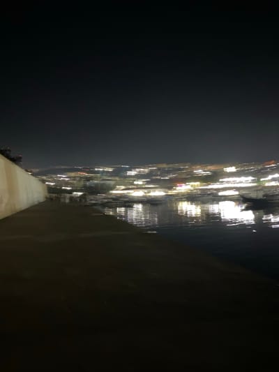A nighttime view of a city skyline across a body of water. The lights of the city are blurred, indicating a long exposure or movement during the photograph. In the foreground, there is a dark, sloped area leading down to the water. To the left, a light-colored wall runs along the edge of the sloped area. The water reflects the city lights in streaks. The overall impression is of a vibrant, illuminated cityscape at night, viewed from a waterfront vantage point. This scene is likely located in Tipaza, Algeria, given the prompt.