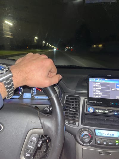 The image depicts the interior of a vehicle at night, specifically in the city of Musachevo, Bulgaria, from the driver's perspective. A male's right hand, wearing a metallic wristwatch with a blue bezel, firmly grips the black steering wheel. The dashboard's instrument cluster is partially visible, showing an odometer reading of 245,660 km and a trip meter of 268.7 km. A Pioneer touchscreen head unit occupies the center console, displaying "8:23" and several radio presets, including "P2=98.00MHz FM" and "P6=JAZZ FM." Below it, a second Pioneer unit is labeled "VERONIKA." Climate controls show a set temperature of 24.0°C in "AUTO" mode. Keys are visible dangling from the ignition. Through the windshield, a dark, wet road is visible, illuminated by a linear array of streetlights on the left, and blurred red and white lights from distant vehicles. The windshield exhibits minor smudges and reflections of interior lights.
