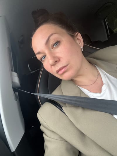 The image displays a headshot of a woman taken inside a car, oriented 90 degrees counter-clockwise. The woman has fair skin, blue eyes, and dark hair pulled back, with a few strands framing her face. She wears a white t-shirt, a light beige or tan blazer, and a thin silver chain necklace. A small, sparkly stud earring is visible in her left ear (from the viewer's perspective). Her expression is neutral to slightly pensive as she looks directly towards the camera.

She is secured by a grey seatbelt with dark stripes, which diagonally crosses her chest. The car interior is predominantly dark, likely black or dark grey, with a leather or faux leather texture visible on the seat or headrest directly behind her. Upside down, the word "AIRBAG" is discernible on this dark surface, indicating a safety feature within the car's interior. Below her, towards the bottom of the frame, there is a lighter grey textured panel, possibly part of the car's dashboard or lower door trim. The lighting appears soft and natural, suggesting an overcast day or perhaps early morning/late afternoon. The scene takes place in a car in Kryukovo, Russia.