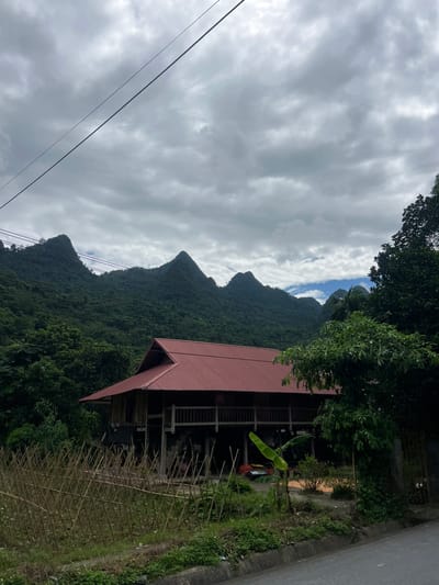 The image captures a scenic view of the countryside in Thuận Hòa Commune, Vietnam, under a cloudy sky. Dominating the background are lush, green mountains with distinctive jagged peaks, partially obscured by a network of power lines. In the foreground, a traditional stilt house with a red metal roof stands prominently. The house is raised on wooden stilts and features a balcony. In front of the house, a rustic bamboo fence encloses a garden area with various plants, including what appears to be a banana tree. A paved road runs along the bottom right of the frame, with a concrete curb. The overall scene depicts a tranquil rural setting, blending natural landscapes with local architecture.