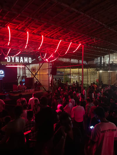 This image captures a lively scene at a nightclub in Vagator, India, illuminated by vibrant red neon lights. The nightclub appears to be an open-air venue with a bamboo-covered roof. A crowd of people is gathered, most of them standing and engaged in conversations or observing the activity on a raised platform. On the platform, a DJ is visible behind a set of DJ equipment, suggesting a musical performance is underway. A large screen displaying the word "DYNAMO" is prominent behind the DJ. The overall atmosphere is one of a bustling night out, with the red lighting creating a dynamic and energetic ambiance.