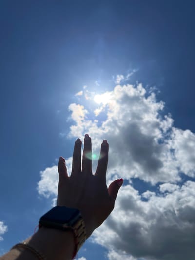 A hand with fingernails painted bright pink is raised against a bright blue sky dotted with white clouds. The sun is visible through the clouds, creating a lens flare effect across the image. A smart watch with a dark screen and a gold-colored band is worn on the wrist. The background is a clear blue sky with scattered clouds, suggesting an outdoor setting in Poland, potentially in the vicinity of Karniowice. The image is taken from a low angle, looking up at the sky.
